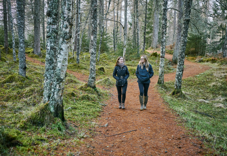 Two young women, dressed in blue jackets, walk together on gravel path through forest of tall fir trees, near Plodda Falls, Glen Affric