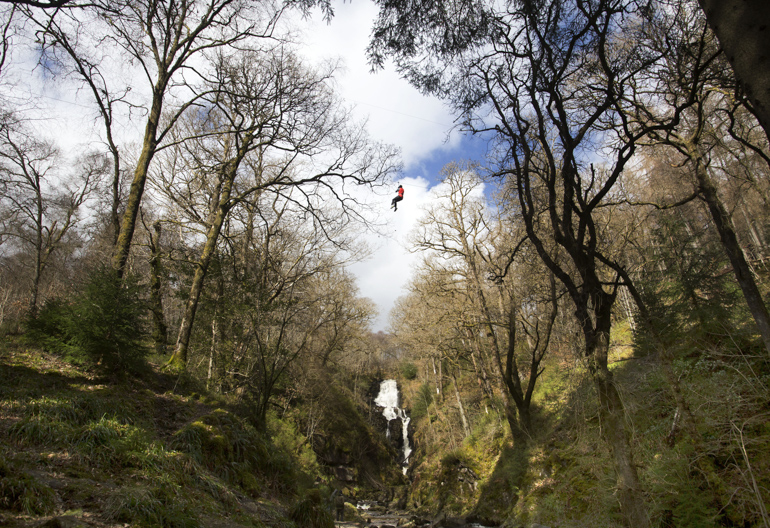 Wide view of person on zip wire across hills beside waterfall, flanked by woodland, Queen Elizabeth Forest Park, Aberfoyle