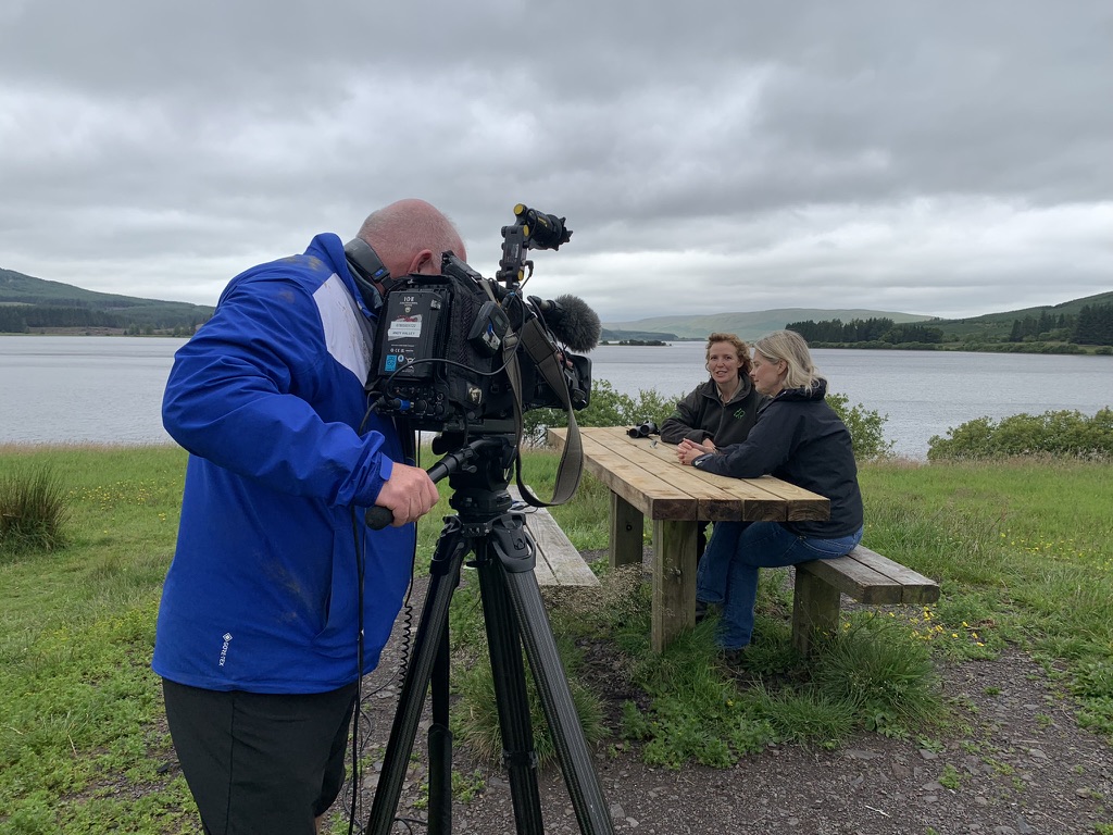 A staff member talking to a camera crew at a picnic table