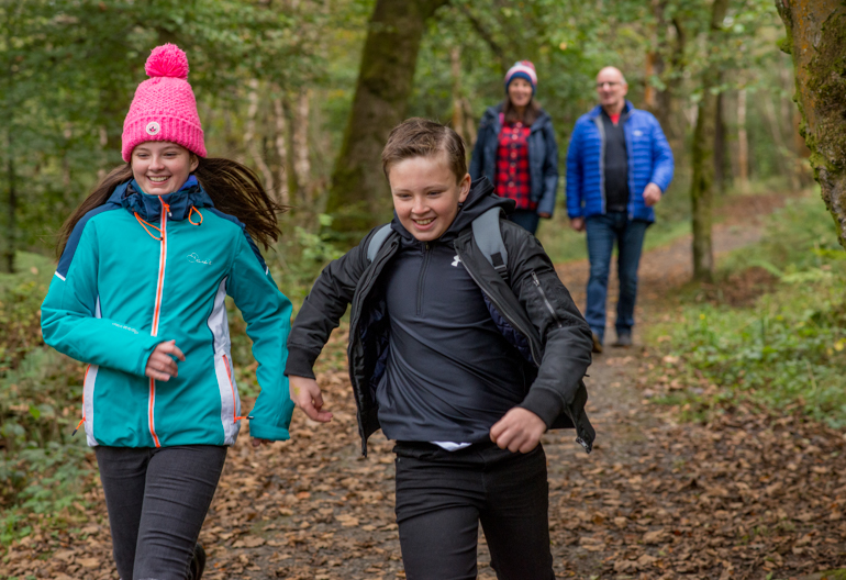 Young girl and boy running together on forest path, followed by man and woman, Bluebell Wood, Renfrewshire Woods, near Johnstone